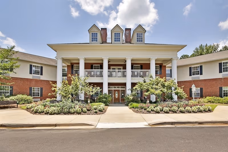 Front exterior view of a large two-story building with white columns, brick and white siding, multiple windows, and a well-maintained garden with shrubs and flowers in front. The building has a central entrance with double doors and a balcony above supported by the columns.