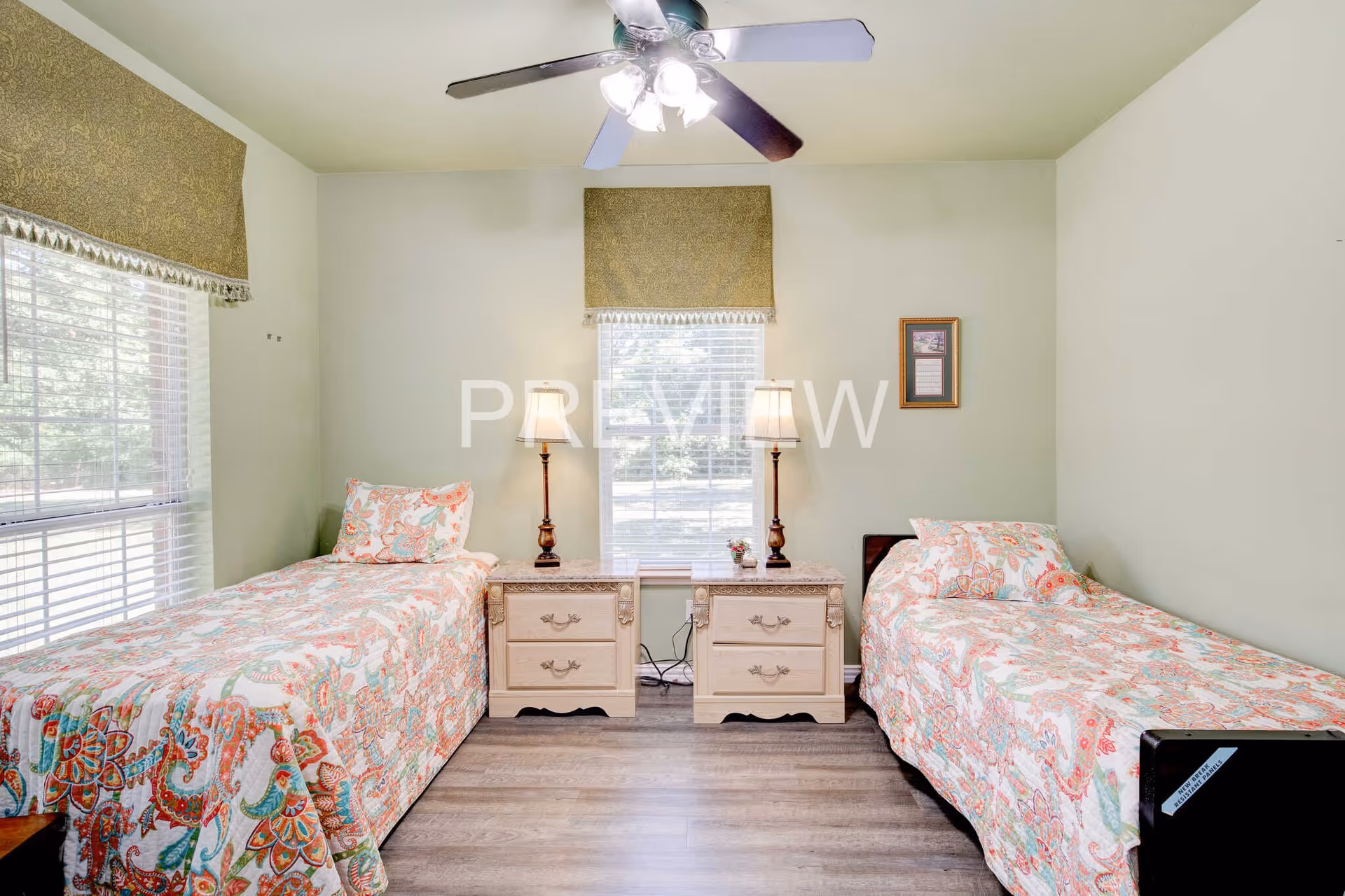 A bedroom with two twin beds placed against opposite walls, each with colorful patterned bedspreads and pillows. Between the beds are two matching nightstands with table lamps and a small decorative item. The room has light green walls, two windows with blinds and green valances, and a ceiling fan with lights. The floor is wood-style laminate.