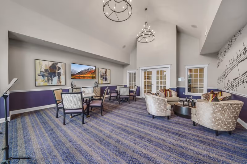 A spacious senior living common area with blue and purple striped carpet, several round tables with chairs, and a seating area with patterned armchairs and a round coffee table. The walls are decorated with musical-themed artwork and a large musical notes mural. Two large chandeliers hang from the high ceiling, and there are double glass doors and windows on one wall.