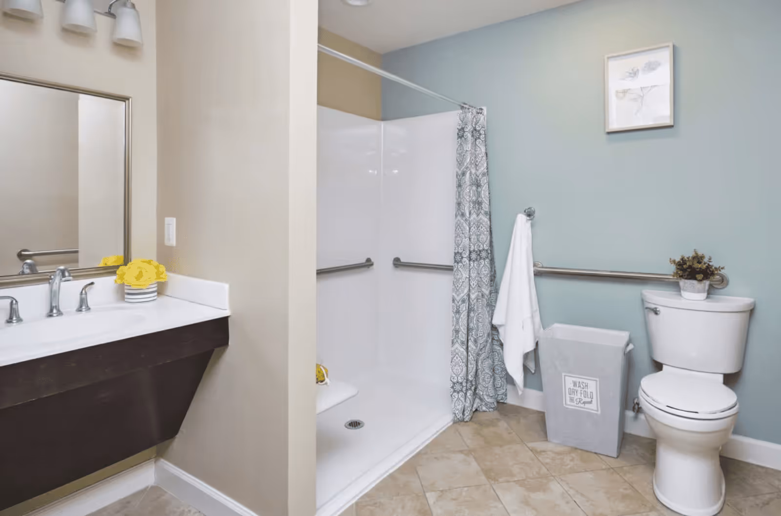 A clean and accessible bathroom featuring a white toilet with a small plant on top, a walk-in shower with grab bars and a patterned shower curtain, a towel hanging on a grab bar, a laundry hamper with the text 'WASH DRY FOLD Repeat', and a sink with a mirror and yellow flowers in a striped vase on the counter.