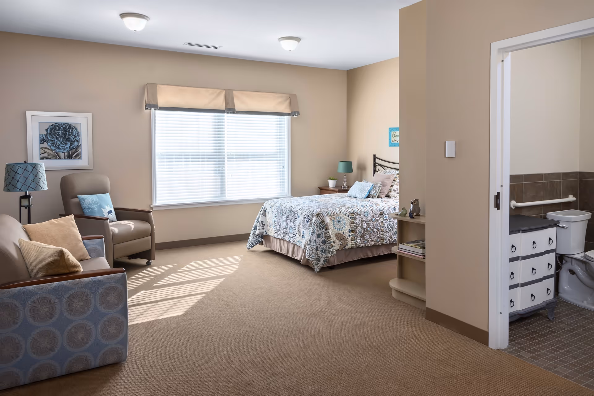 A bright and spacious senior living bedroom with a bed covered in a patterned quilt, a nightstand with a lamp and plant, a comfortable armchair, a sofa with pillows, a framed flower picture on the wall, and a window with blinds letting in natural light. To the right, there is an open doorway leading to a bathroom with a toilet and a vanity with drawers.