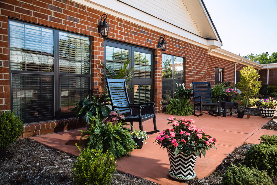 Outdoor patio area with black rocking chairs and various potted plants and flowers in front of a brick building with large windows and two wall-mounted lantern lights.