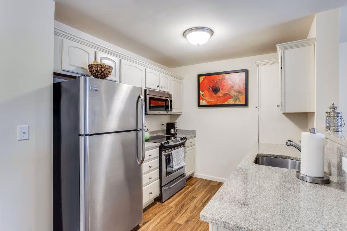 A modern kitchen with white cabinets, stainless steel refrigerator, stove, and microwave. The kitchen has granite countertops, a sink with a paper towel holder, and a coffee maker. A colorful painting of red flowers hangs on the wall.