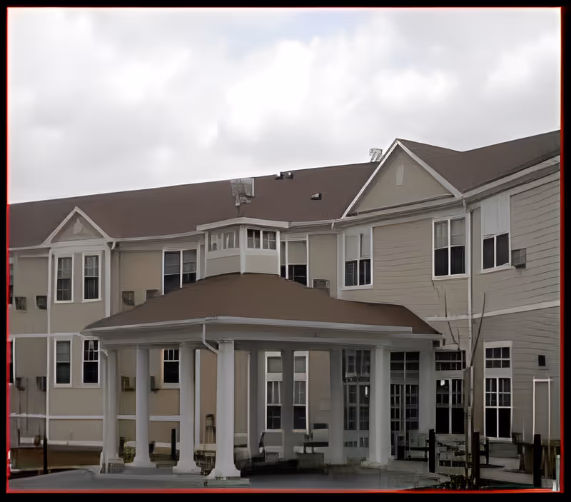 Exterior view of a multi-story assisted living facility building with beige siding and white trim. The building features a covered entrance supported by white columns and multiple windows. The sky is cloudy.