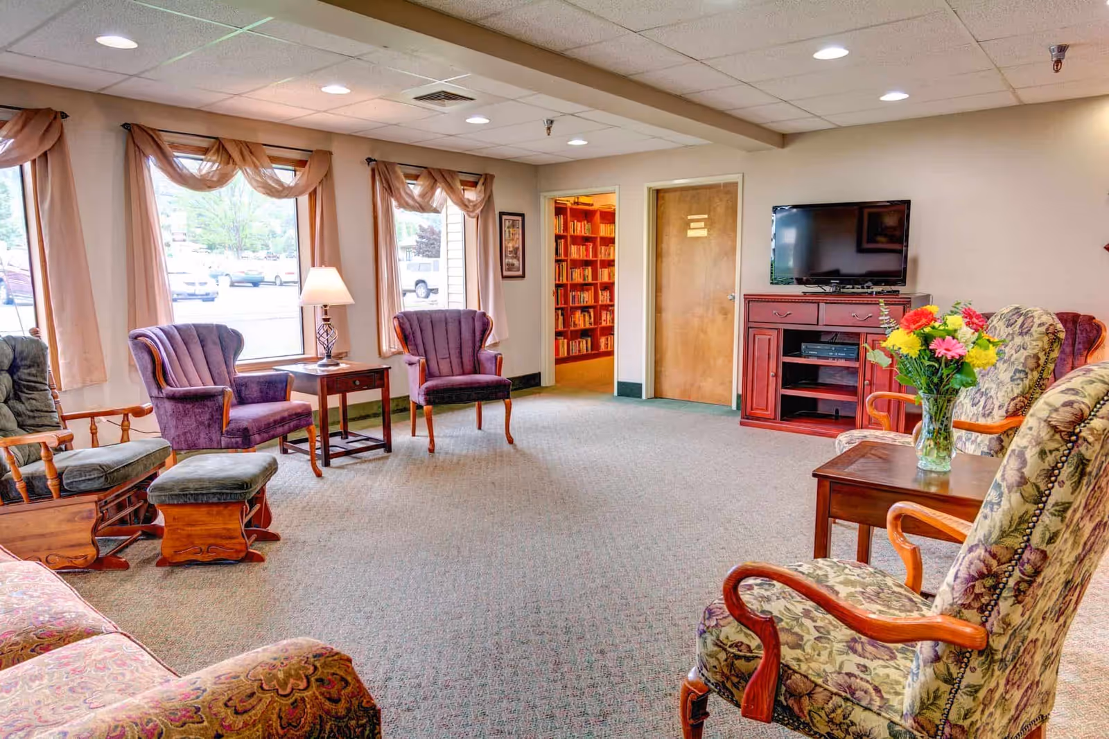 A bright and cozy living room area in a retirement residence with several upholstered armchairs and a wooden rocking chair with a matching footstool. There is a wooden side table with a lamp between two purple chairs near large windows with sheer curtains. A flat-screen TV is mounted on a red wooden cabinet, and a vase with colorful flowers sits on a small table next to floral-patterned chairs. A doorway leads to a room filled with bookshelves.