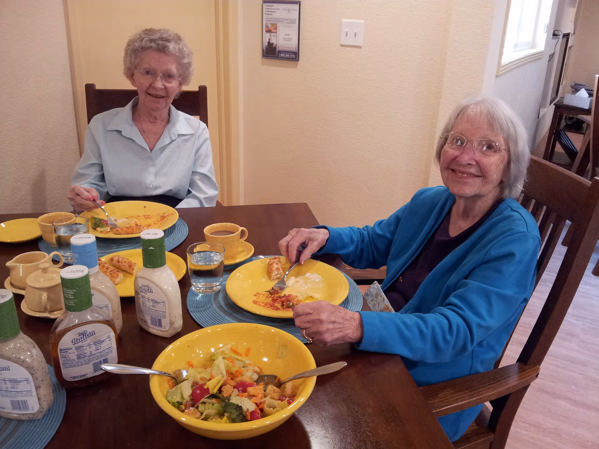 Two elderly women sitting at a dining table in a senior living facility, eating a meal. The table has yellow plates, cups, glasses of water, salad in a yellow bowl, and several bottles of salad dressing. The women are smiling and appear to be enjoying their meal in a cozy indoor setting.