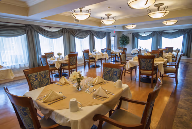 A spacious dining room with multiple tables set for a meal. Each table is covered with a white tablecloth and a beige table runner, with neatly folded beige napkins, glassware, and silverware. The chairs have wooden frames with patterned upholstery. Large windows with blue curtains allow natural light to fill the room, and ceiling lights provide additional illumination.