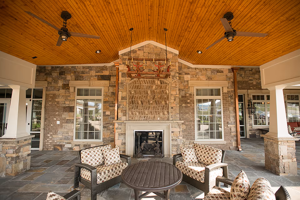Covered outdoor seating area with stone walls and a fireplace in the center. Four cushioned wicker chairs with patterned pillows surround a round wooden table. The ceiling is wooden with two ceiling fans and a rustic chandelier hanging above the fireplace. Large windows and columns frame the space.