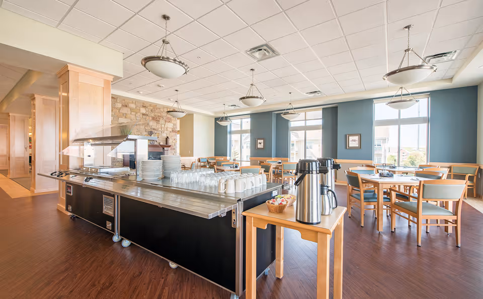 A bright and spacious dining area in a senior living facility with multiple wooden tables and chairs arranged neatly. Large windows allow natural light to fill the room. In the foreground, there is a serving station with stacks of plates, glasses, and mugs, along with a small table holding two large coffee dispensers and a basket of creamers.