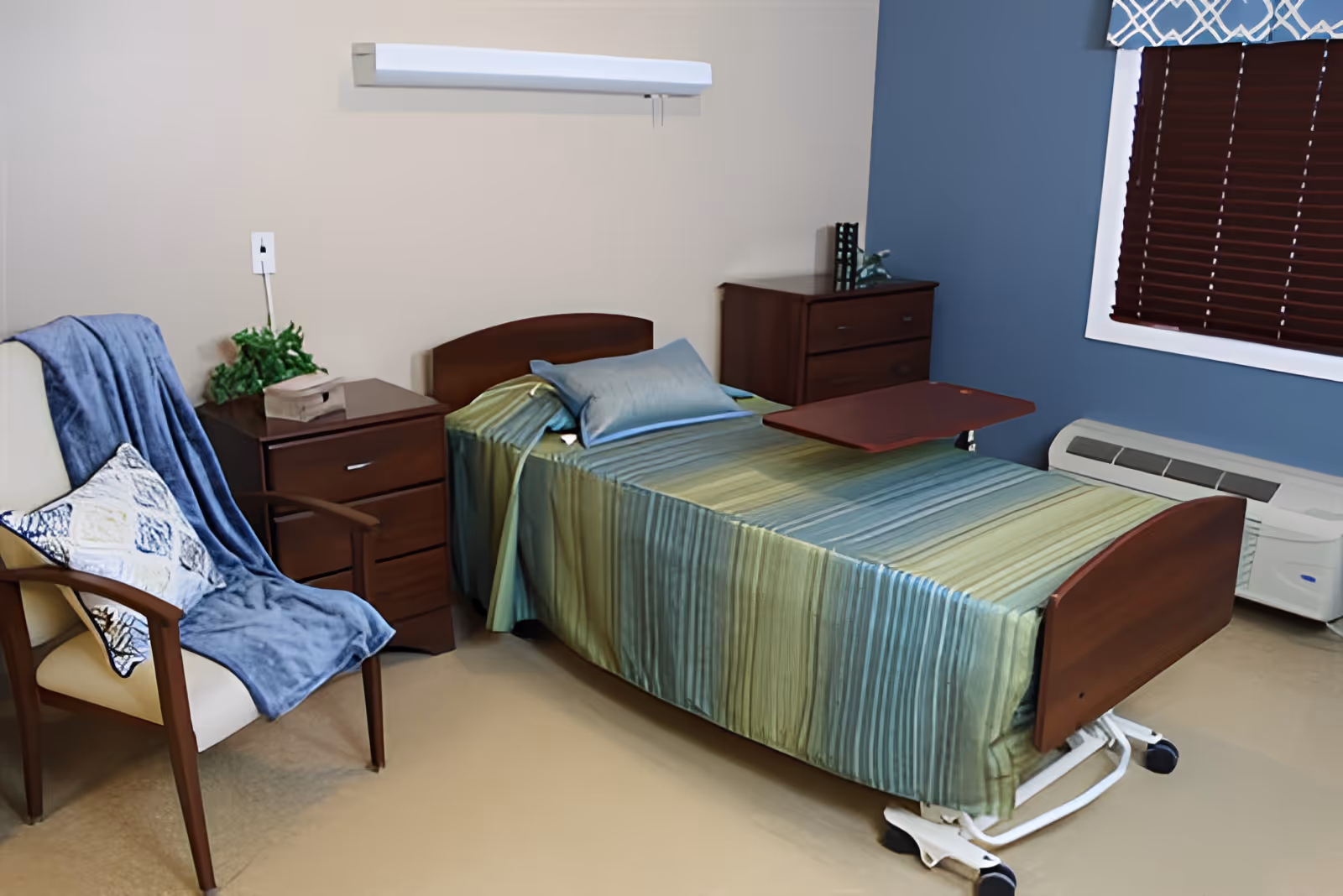 A small bedroom in a senior living facility featuring a single hospital-style bed with a green and blue striped bedspread and a blue pillow. Next to the bed is a wooden nightstand with a plant and a tissue box. A wooden chair with a blue throw blanket and a decorative pillow is positioned beside the nightstand. The room has beige walls with one blue accent wall, a window with dark blinds and a patterned valance, and a white air conditioning unit below the window.