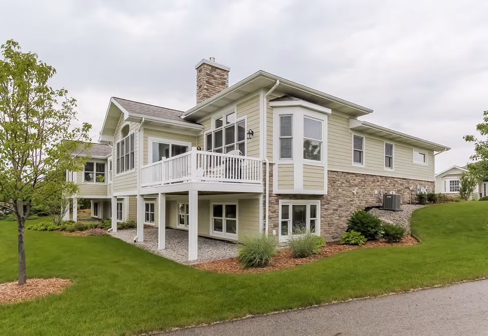 Exterior view of a two-story residential building with beige siding and stone accents, featuring a white balcony and surrounded by green grass and small trees under a cloudy sky.
