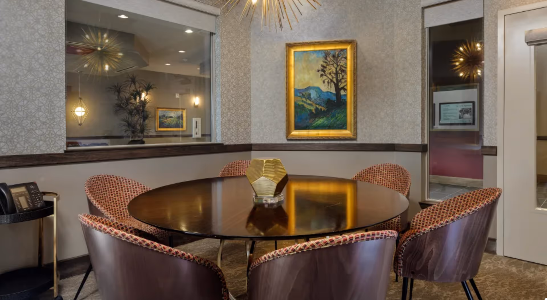 A small meeting or dining area with a round wooden table surrounded by six upholstered chairs with patterned fabric. A decorative golden geometric centerpiece sits on the table. The walls are decorated with patterned wallpaper and a framed landscape painting. There is a window looking into another room and a door with a glass panel. A telephone is placed on a small side table to the left.
