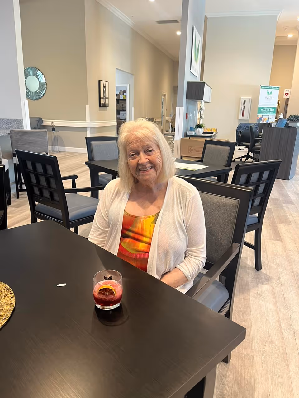 An elderly woman smiling while seated at a table with a drink in a communal dining area of a senior living facility.