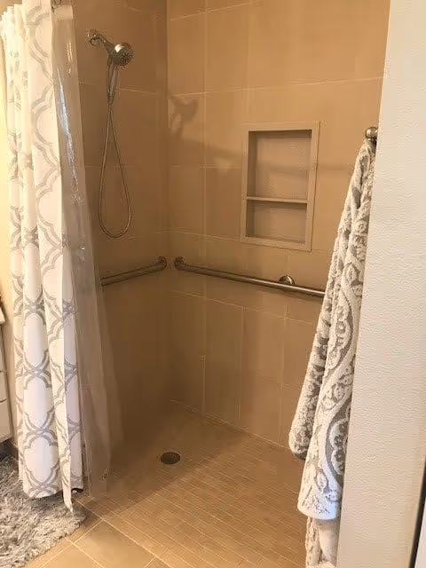 A tiled walk-in shower with beige tiles, a handheld showerhead, and two horizontal grab bars on the walls. There is a built-in recessed shelf for toiletries and a patterned shower curtain partially drawn to the side. A patterned towel hangs on a hook outside the shower.