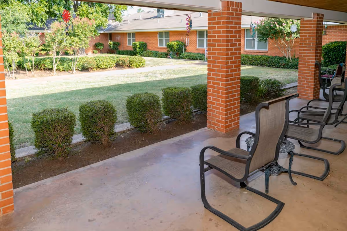 Covered outdoor patio area with several metal-framed chairs with mesh seats and backs, brick pillars, and a view of a grassy courtyard with trimmed bushes and a brick building in the background.