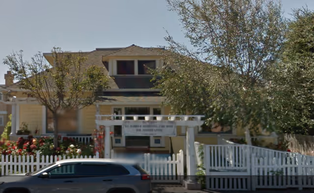 Front exterior view of Bay Harbour Care Home, a yellow residential-style building with a white picket fence, trees, and a parked car in front.