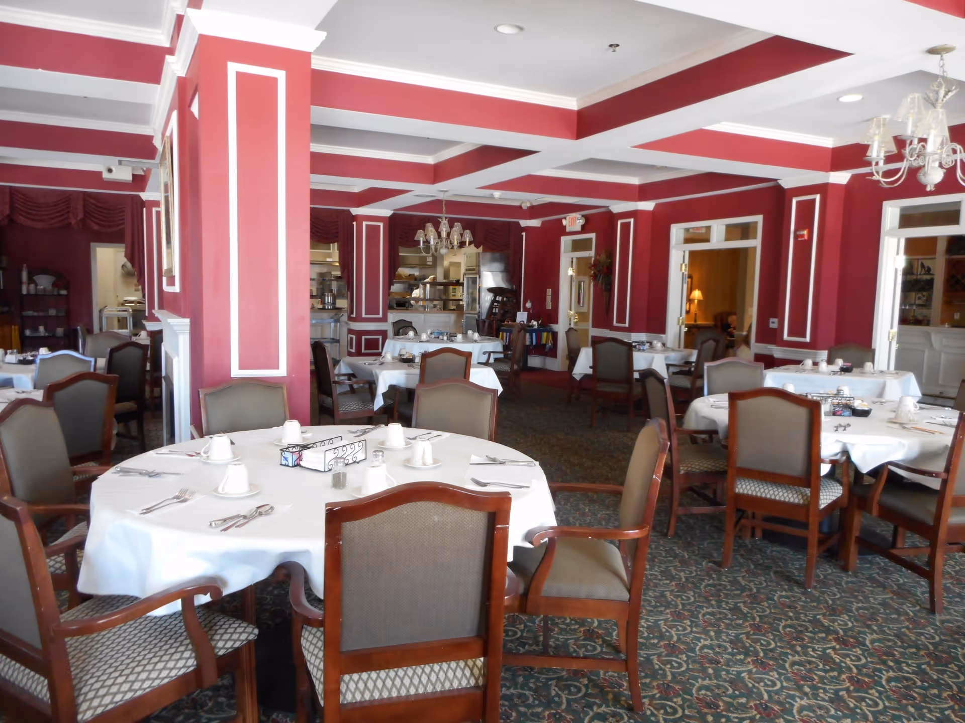 Dining room with round tables covered in white tablecloths, set with cups, utensils, and napkins. The room has red walls with white trim, chandeliers hanging from the ceiling, and patterned carpet flooring. Several wooden chairs with cushioned seats surround the tables. The background shows an open kitchen area and doorways leading to other rooms.