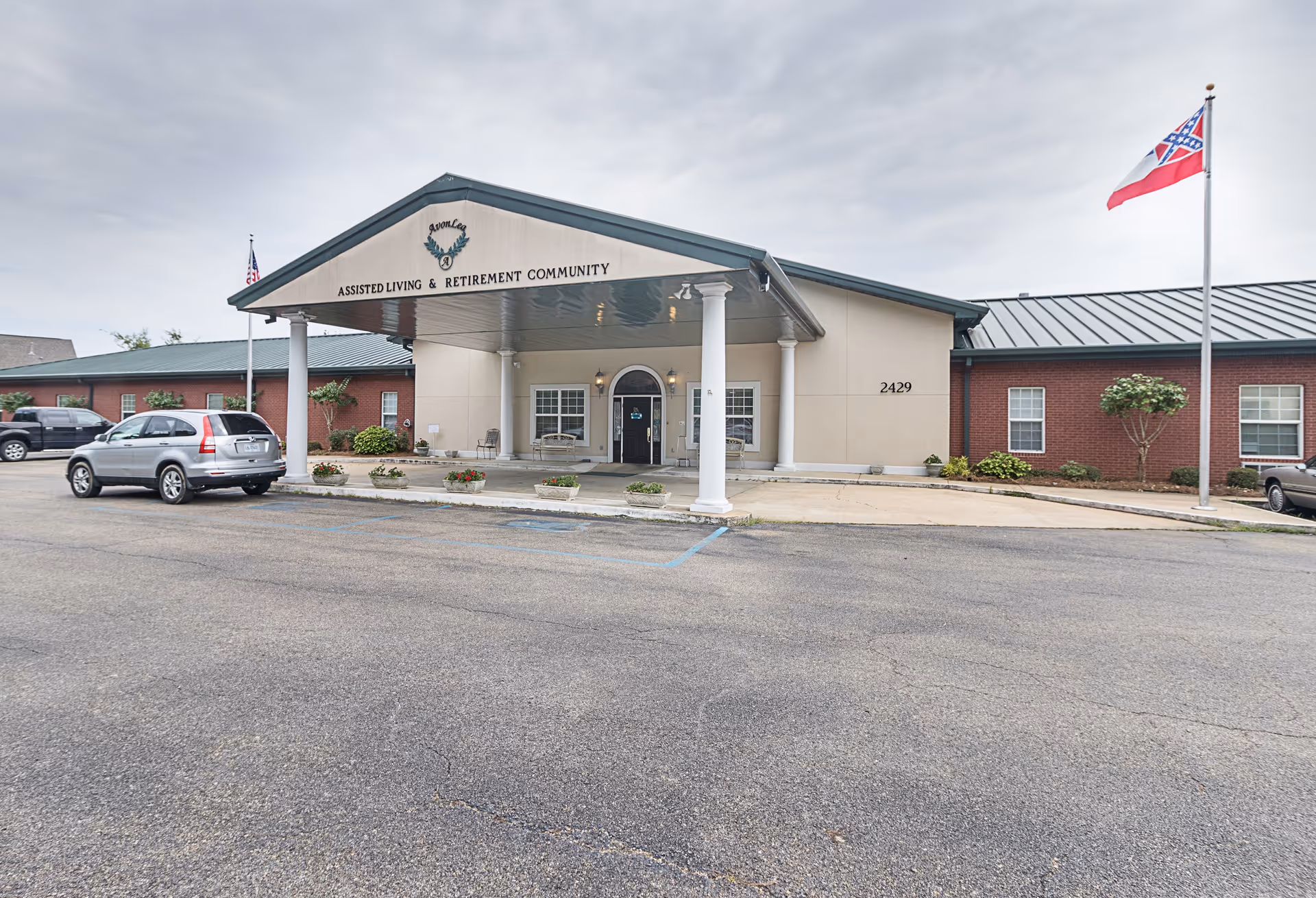 Entrance of an assisted living and retirement community building with a covered portico, columns, flagpoles, and parked cars.
