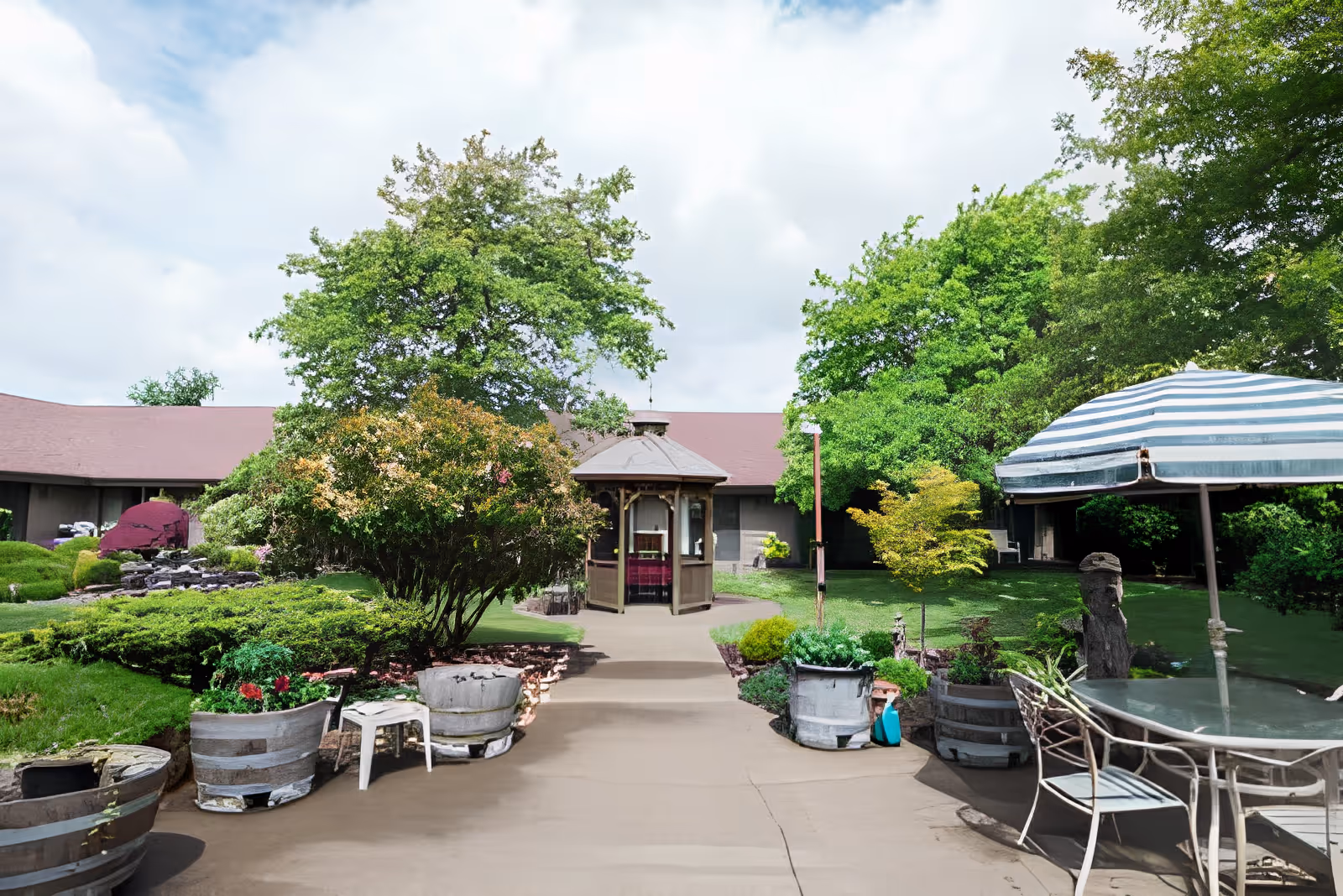 Sunny courtyard with a central gazebo, potted plants, patio table and umbrella, and a surrounding single-story building.