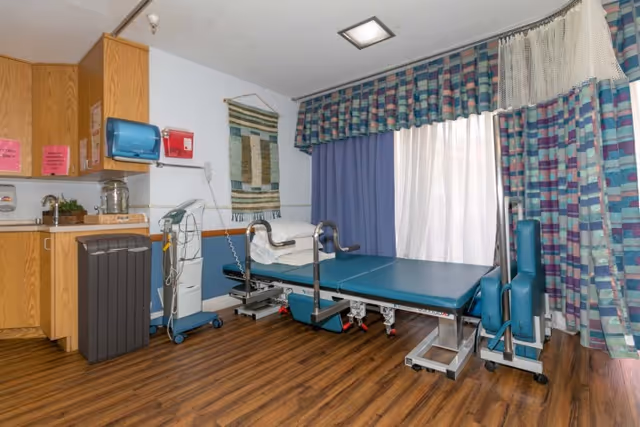 Medical treatment/therapy room with a blue adjustable exam/therapy table, wooden cabinets and sink, and patterned curtains over a window.