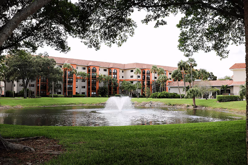 Multi-story assisted living building viewed across a pond with a central fountain, framed by trees and lawn.