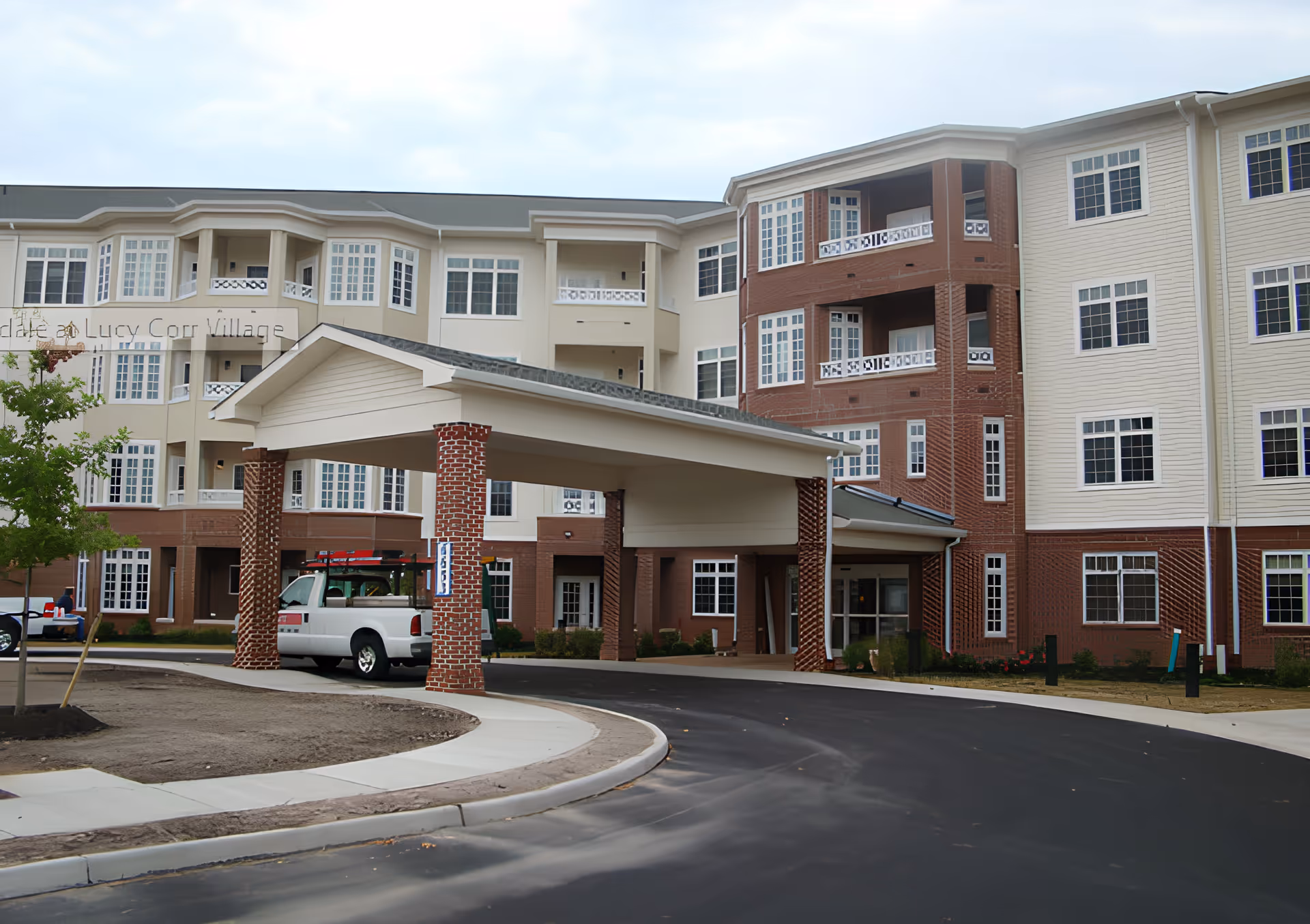 Exterior view of The Assisted Living at Lucy Corr Village building with a covered entrance supported by brick columns, a white pickup truck parked under the entrance, and a curved driveway leading to the entrance. The building has multiple windows and a mix of beige and brick facade.