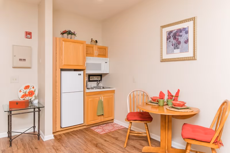 Small kitchen area with wooden cabinets, a white microwave, and a refrigerator. A round wooden dining table with two wooden chairs with red cushions is set with red napkins and green cups. A framed floral picture hangs on the wall above the table. There is a small glass side table with a decorative plate and a red box on it. The floor is wooden, and the walls are light-colored.