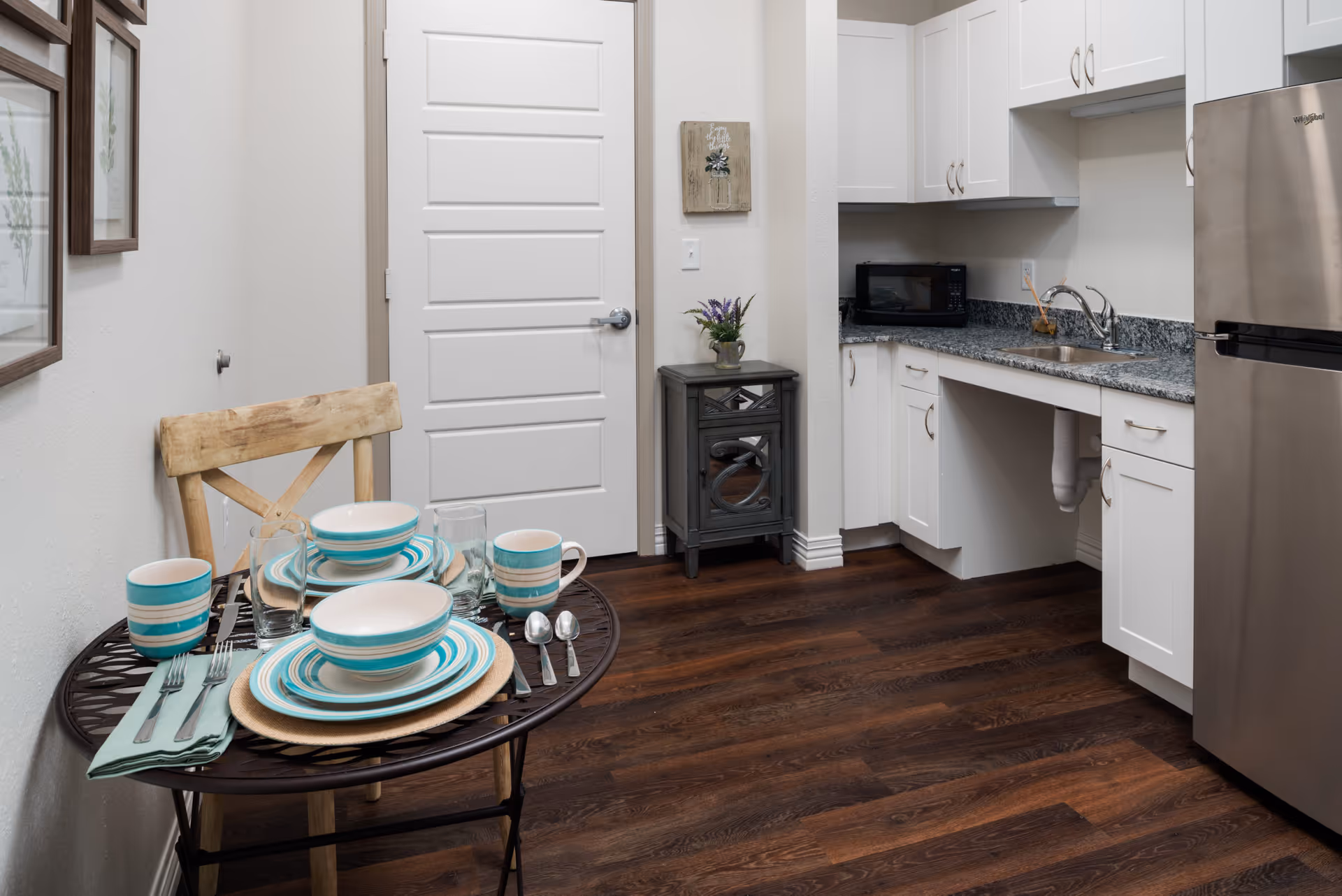 Small kitchenette with a set dining table and wooden chair, white cabinets, sink and stainless-steel refrigerator.
