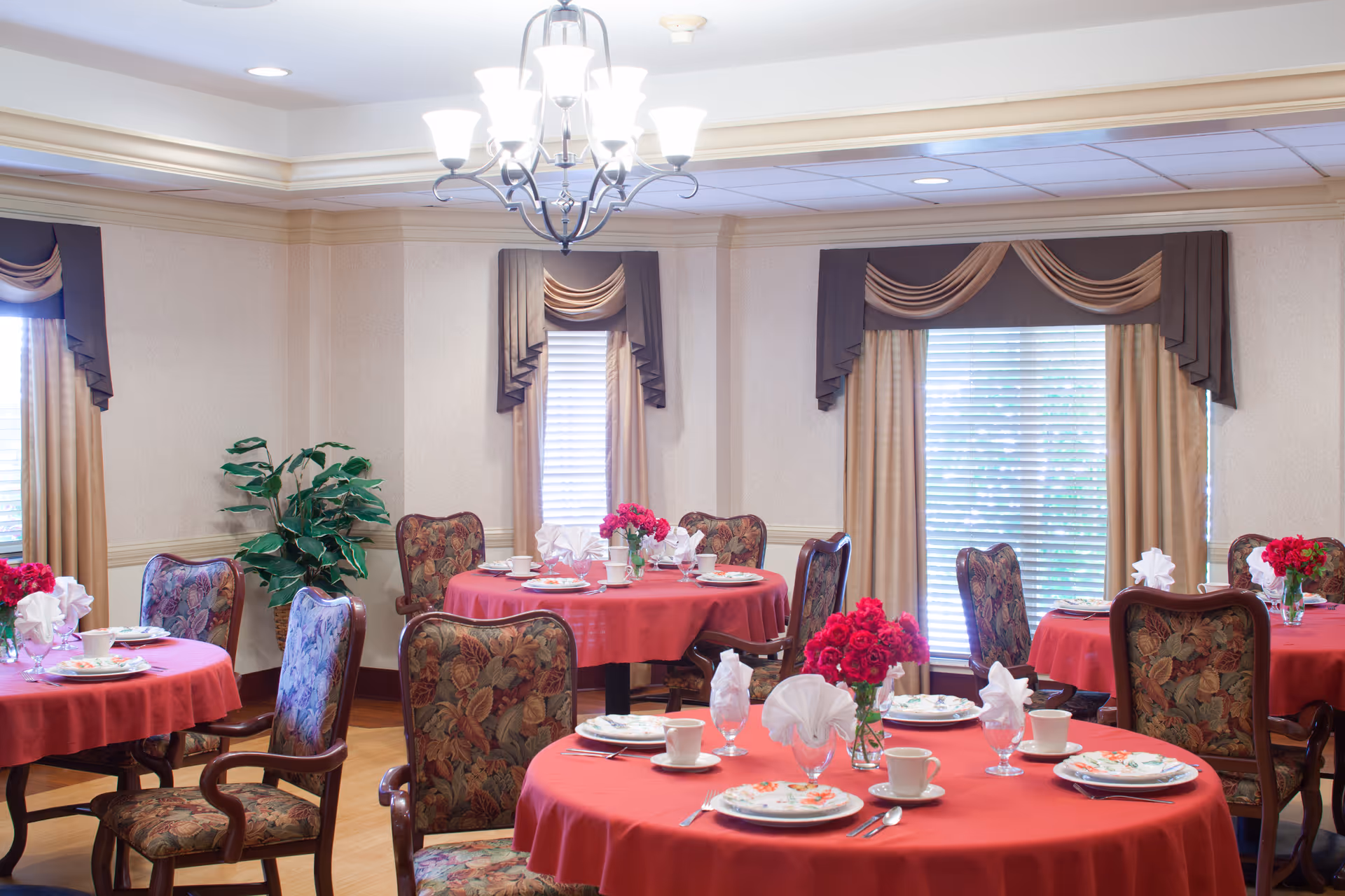 Decorative dining room with round tables draped in red tablecloths, place settings and floral centerpieces beneath a chandelier.