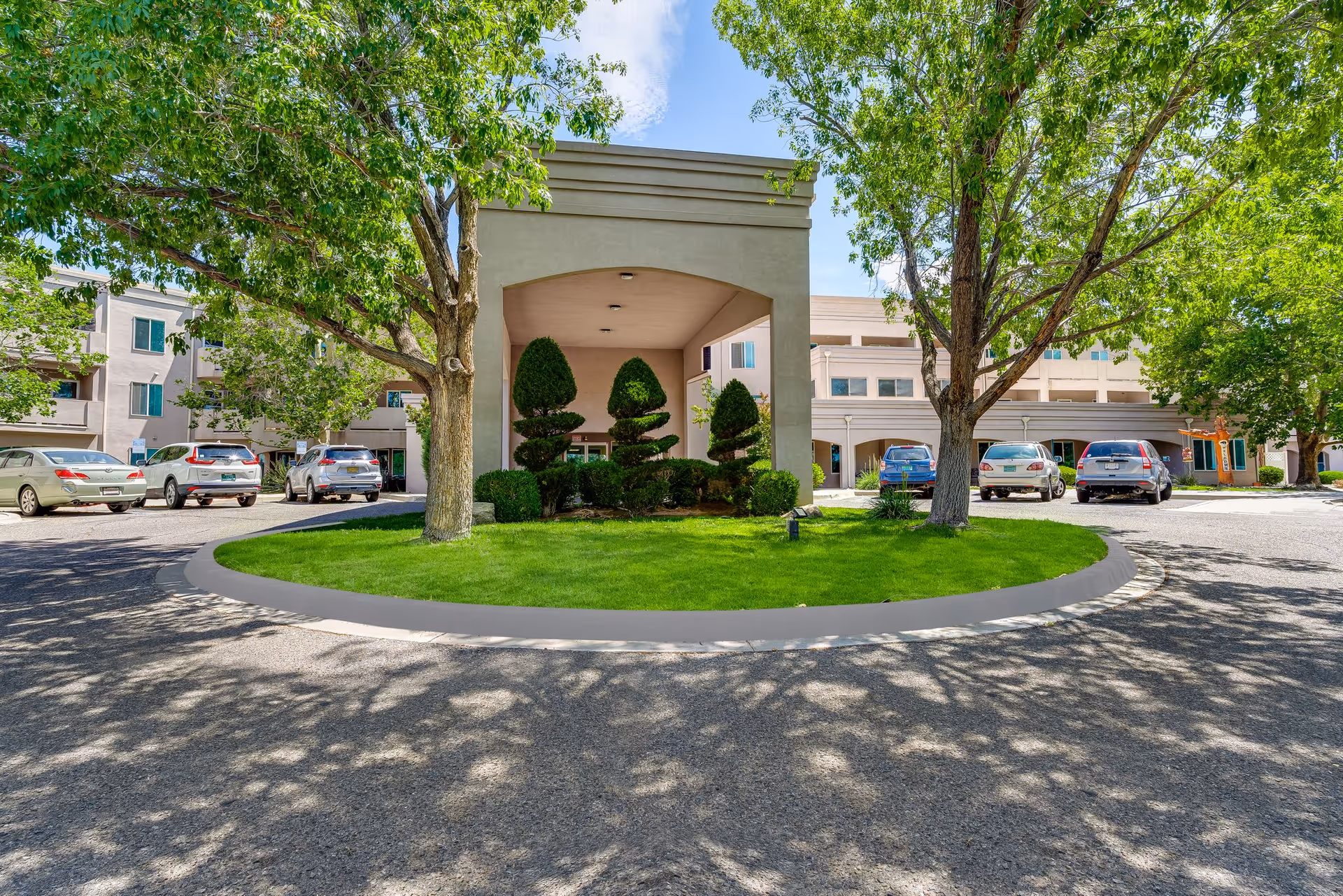Front exterior view of Bear Canyon Estates | Sky Active Living facility showing a circular driveway with green grass and sculpted bushes in the center, flanked by trees and parked cars, under a clear blue sky.