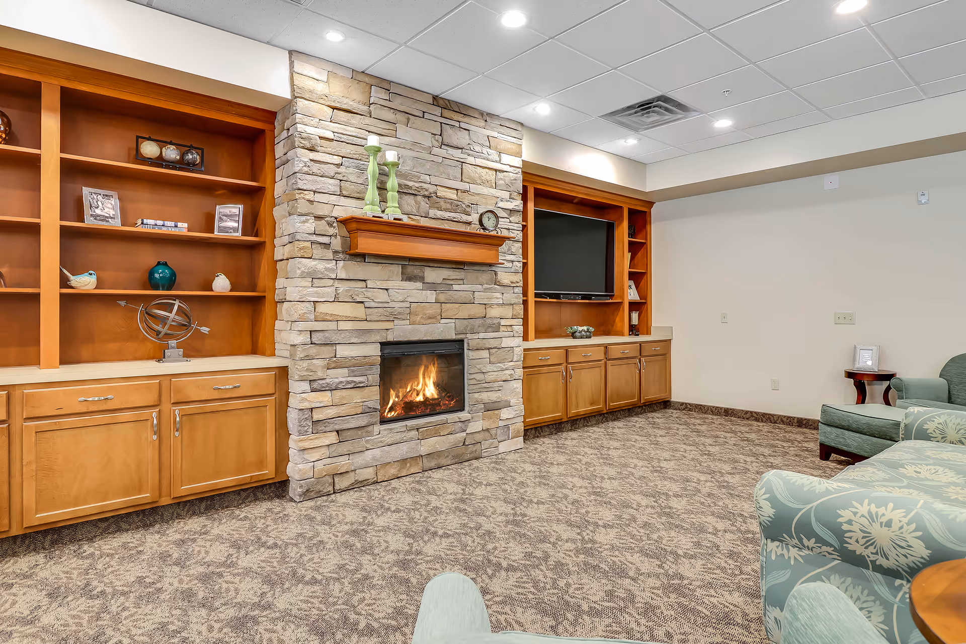 A cozy living room area featuring a stone fireplace with a wooden mantel, built-in wooden shelves and cabinets on either side, a flat-screen TV on the right side, and comfortable upholstered chairs with floral patterns. The room has a carpeted floor and recessed ceiling lights.