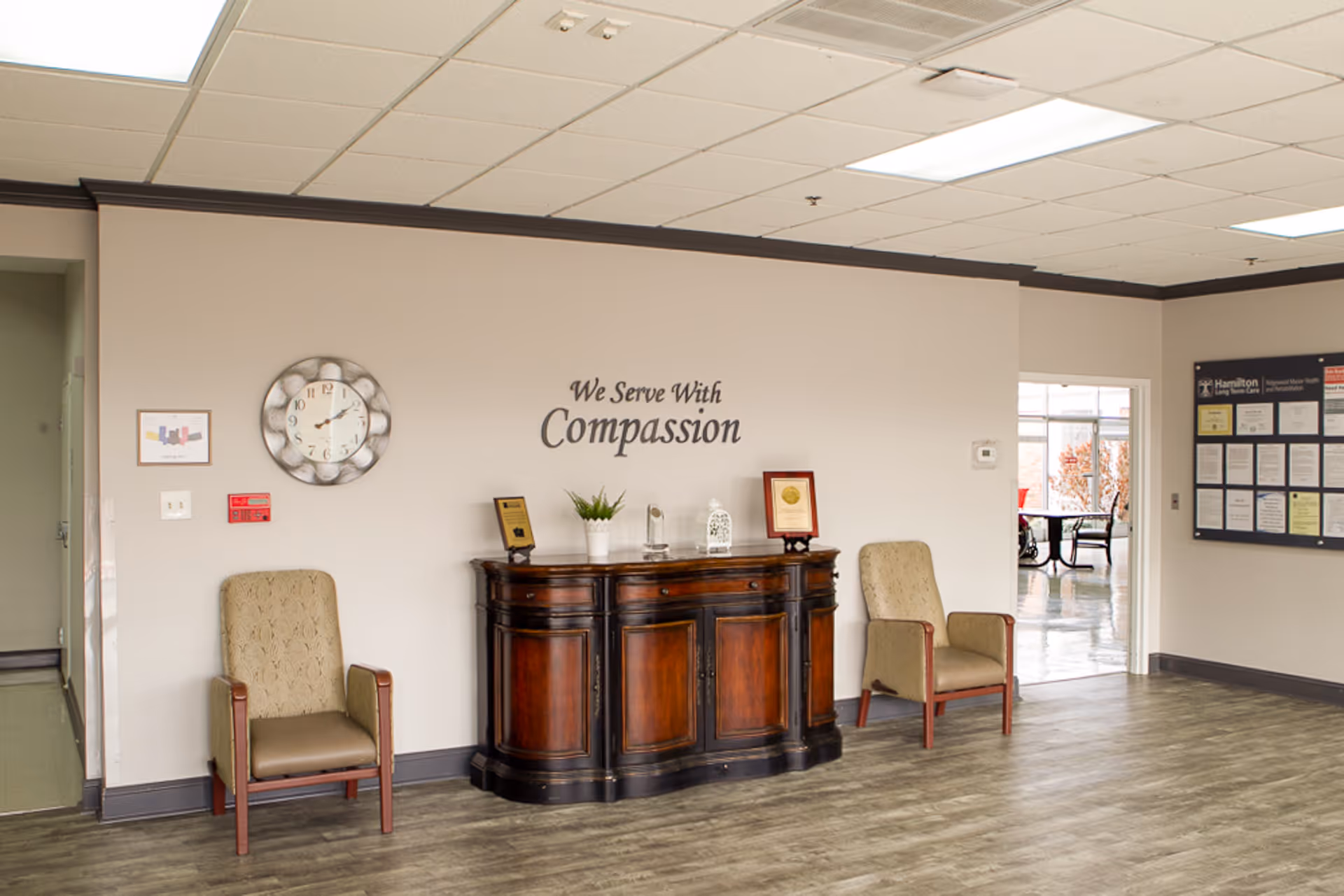 A waiting area in a senior living facility with two beige armchairs on either side of a dark wooden cabinet. Above the cabinet, the wall has the phrase 'We Serve With Compassion'. A round wall clock and a framed picture are on the left wall. On the right wall, there is a bulletin board with various notices. The floor is wood-patterned, and there is a doorway leading to another room with tables and chairs.
