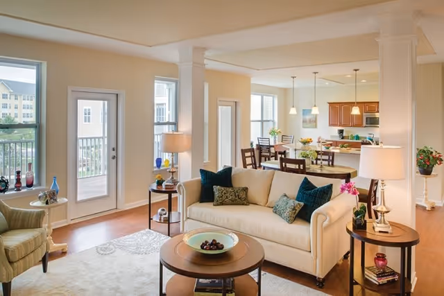Bright and spacious living room with a cream-colored sofa adorned with blue and patterned cushions, two side tables with lamps and decorative items, a round coffee table with a bowl of fruit, an armchair, and large windows and a glass door leading to a balcony. In the background, a dining area with a wooden table and chairs, pendant lights, and a kitchen with wooden cabinets and stainless steel appliances are visible.