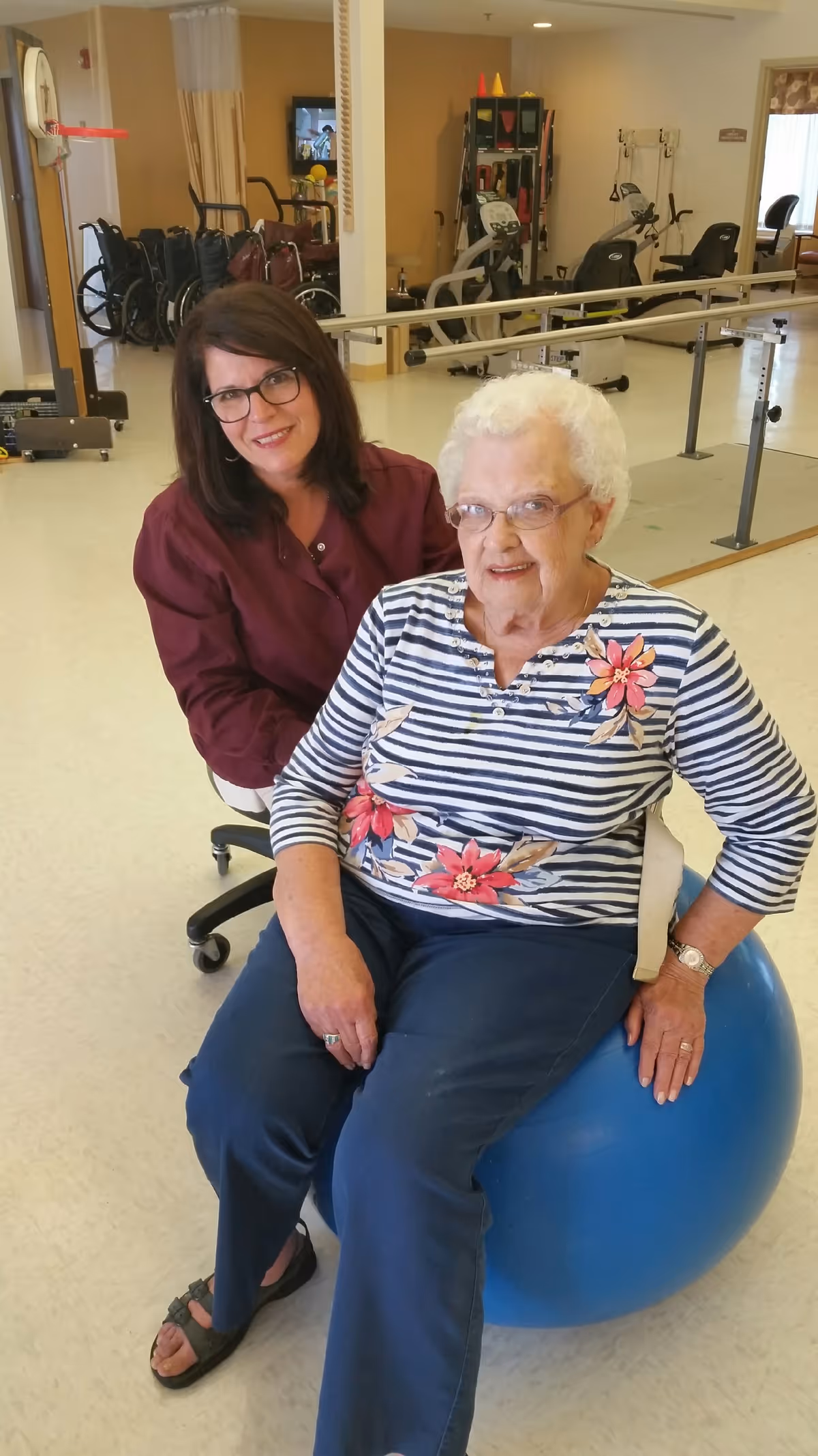An elderly woman sitting on a blue exercise ball with a younger woman kneeling beside her in a physical therapy or rehabilitation room. The room contains exercise equipment, wheelchairs, and parallel bars for walking support.