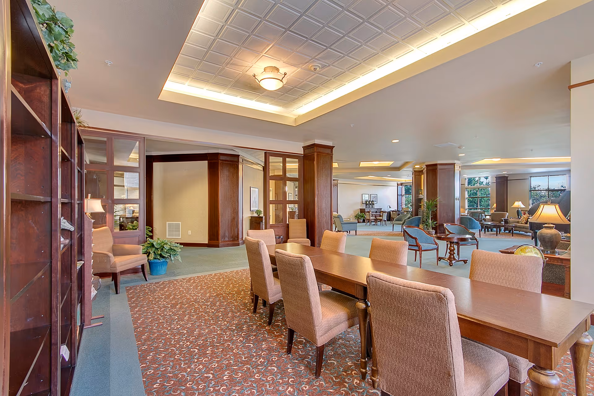 A spacious and well-lit common area in a senior living facility featuring a long wooden table with upholstered chairs, a patterned carpet, wooden columns, large windows, and various seating arrangements including armchairs and small tables. The ceiling has recessed lighting and a decorative light fixture.
