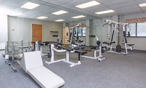 A fitness room with various exercise equipment including weight machines, a bench press, and a padded incline bench. The room has gray carpet, white walls, fluorescent ceiling lights, and a window with floral curtains.