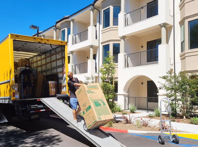 Two men unloading a large U-Haul moving box from a yellow moving truck using a ramp outside a multi-story residential building with balconies and landscaped surroundings.