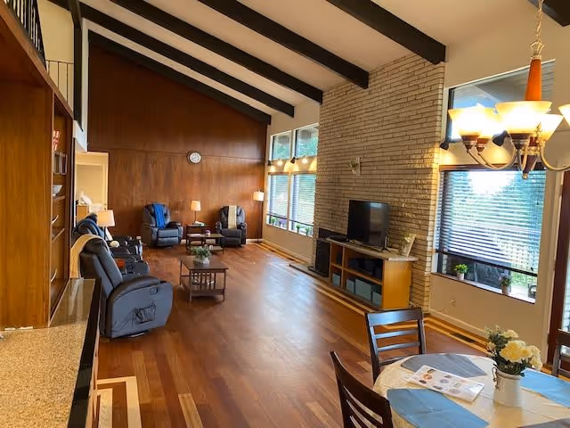 A spacious living room with wooden floors and a high ceiling with exposed dark beams. The room features several recliner chairs arranged around a coffee table, a TV on a wooden stand against a brick wall, and large windows letting in natural light. A dining table with chairs and a chandelier is visible in the foreground, along with some decorative plants and a magazine on the table.