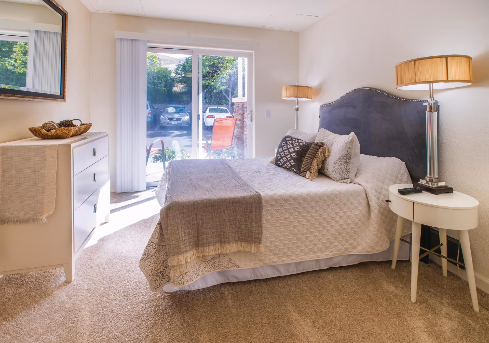 Sunlit bedroom with a neatly made bed, bedside table and lamps, dresser, and sliding glass door to the outside.