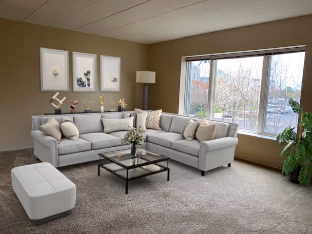 A bright living room with a large L-shaped light gray sofa adorned with several beige and gray cushions. A glass coffee table with a black frame sits in front of the sofa, holding a small vase with flowers. Behind the sofa is a tall floor lamp and three framed abstract art pieces on the wall. Large windows on the right side let in natural light and show a view of trees and buildings outside. A green potted plant is placed near the window.