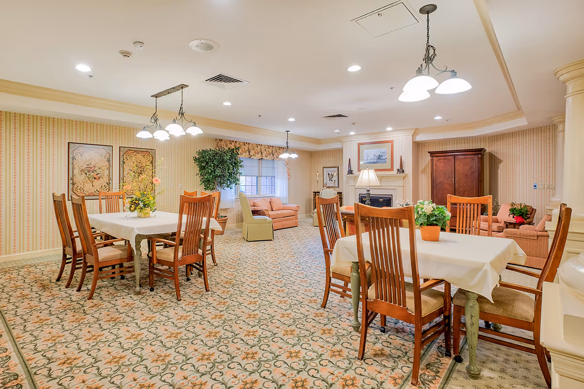 Well-lit senior living dining room and sitting area with multiple tables, wooden chairs, patterned carpet, and a fireplace.