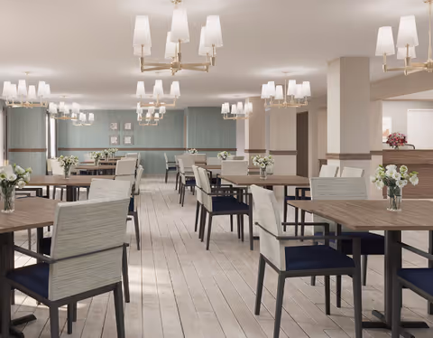 A spacious dining room with multiple wooden tables and cushioned chairs arranged neatly. Each table has a small vase with white flowers. The room features light-colored wooden flooring, soft blue walls, and elegant chandeliers with white lampshades hanging from the ceiling. There is a reception or serving counter visible in the background with a floral arrangement on it.