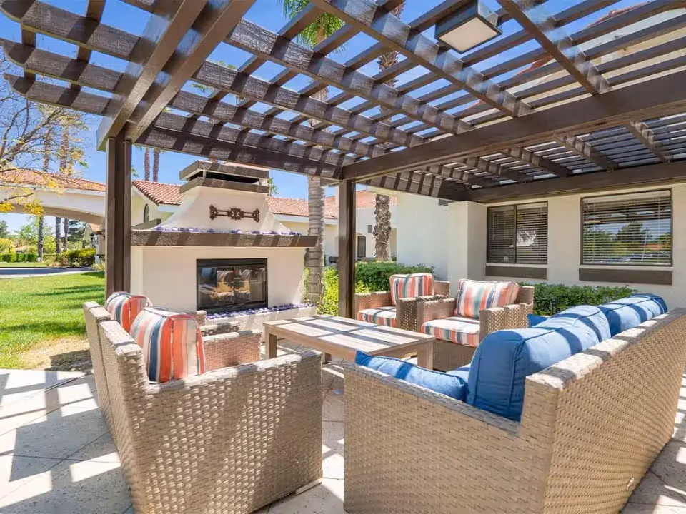 Covered outdoor patio with wicker sofas and armchairs, striped cushions, a central coffee table, and a fireplace beneath a wooden pergola.