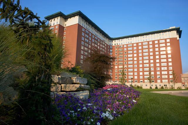 Exterior view of a large multi-story senior living facility building with red and white facade, surrounded by green grass, colorful purple and white flowers, and landscaping with stone steps and trees under a clear blue sky.