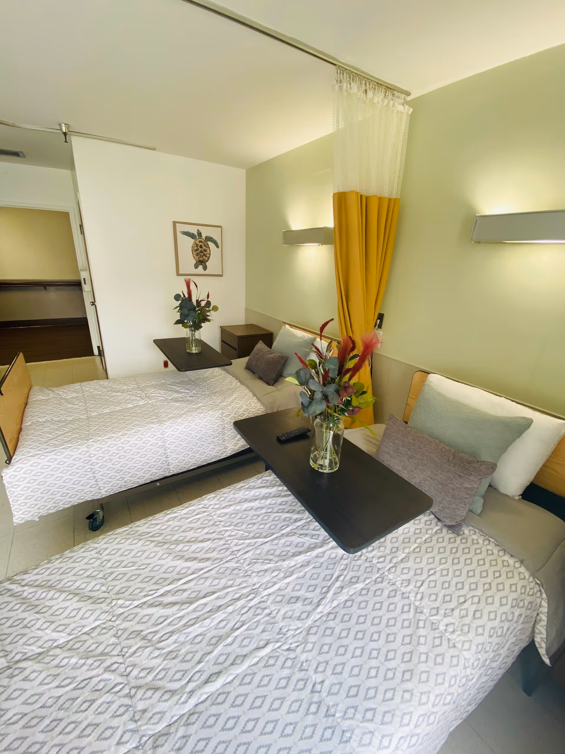 A bright and clean room with two hospital-style beds, each with a patterned white bedspread, pillows, and a black overbed table holding a vase with flowers. A yellow privacy curtain hangs between the beds. The walls are light-colored with modern wall-mounted lights, and a framed picture of a turtle is on the far wall.