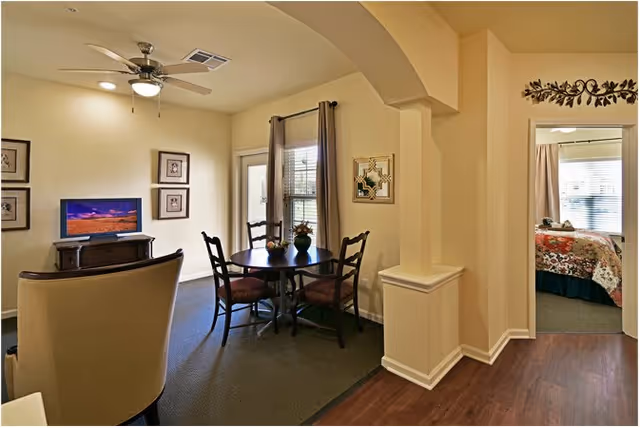 Interior view of a senior living community apartment showing a small dining area with a round table and four chairs, a living area with a TV on a stand, and a glimpse into a bedroom with a bed covered in a floral quilt. The walls are painted beige, and there is a ceiling fan with a light fixture above the dining area. A window with curtains is visible near the dining table.