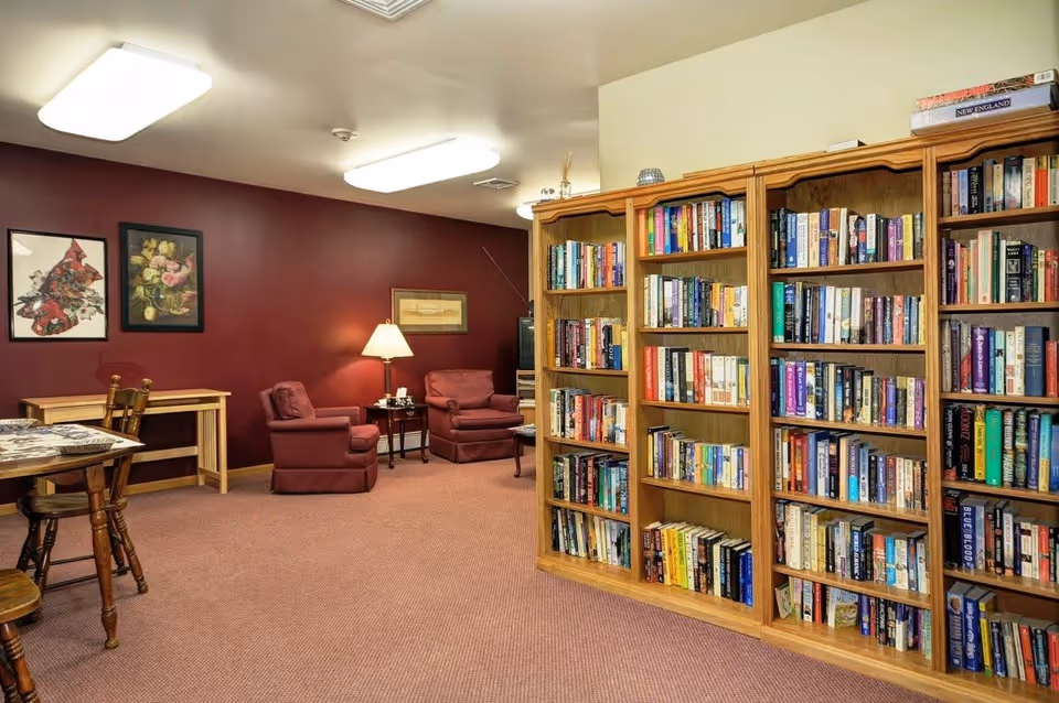 A cozy reading room with wooden bookshelves filled with books on the right side. Two maroon armchairs with a small table and lamp between them are placed against a maroon wall with framed artwork. A wooden table and chairs are on the left side of the room, and the floor is carpeted in a matching maroon tone.