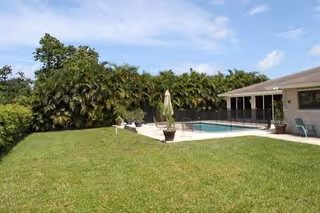 Outdoor view of a grassy yard with a fenced swimming pool surrounded by palm trees and a building with a covered patio area under a partly cloudy sky.
