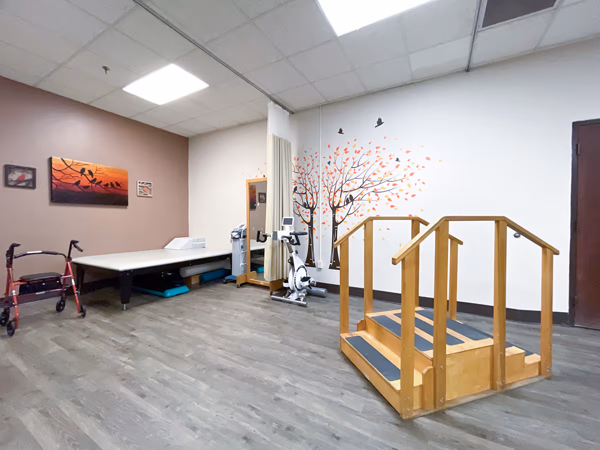A therapy or rehabilitation room with a wooden set of stairs with handrails, an exercise bike, a padded therapy table, a walker, and a wall decorated with a tree and birds mural. The room has a wood floor and a drop ceiling with fluorescent lighting.