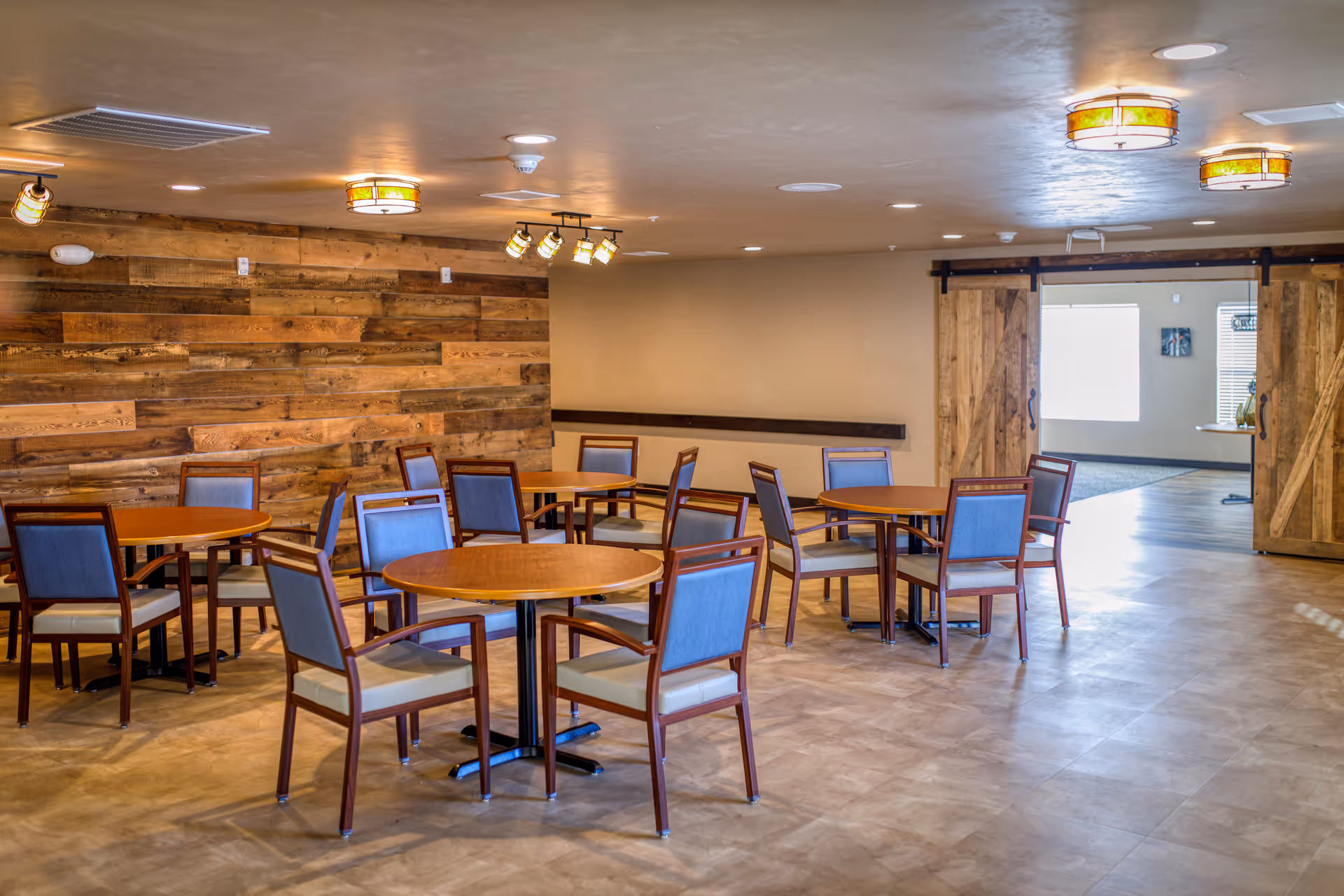 A dining area with several round wooden tables and chairs with blue cushions. The room features a wooden accent wall, beige walls, and a light-colored floor. There are ceiling lights and track lighting, and a set of wooden sliding barn doors leading to another room with windows.